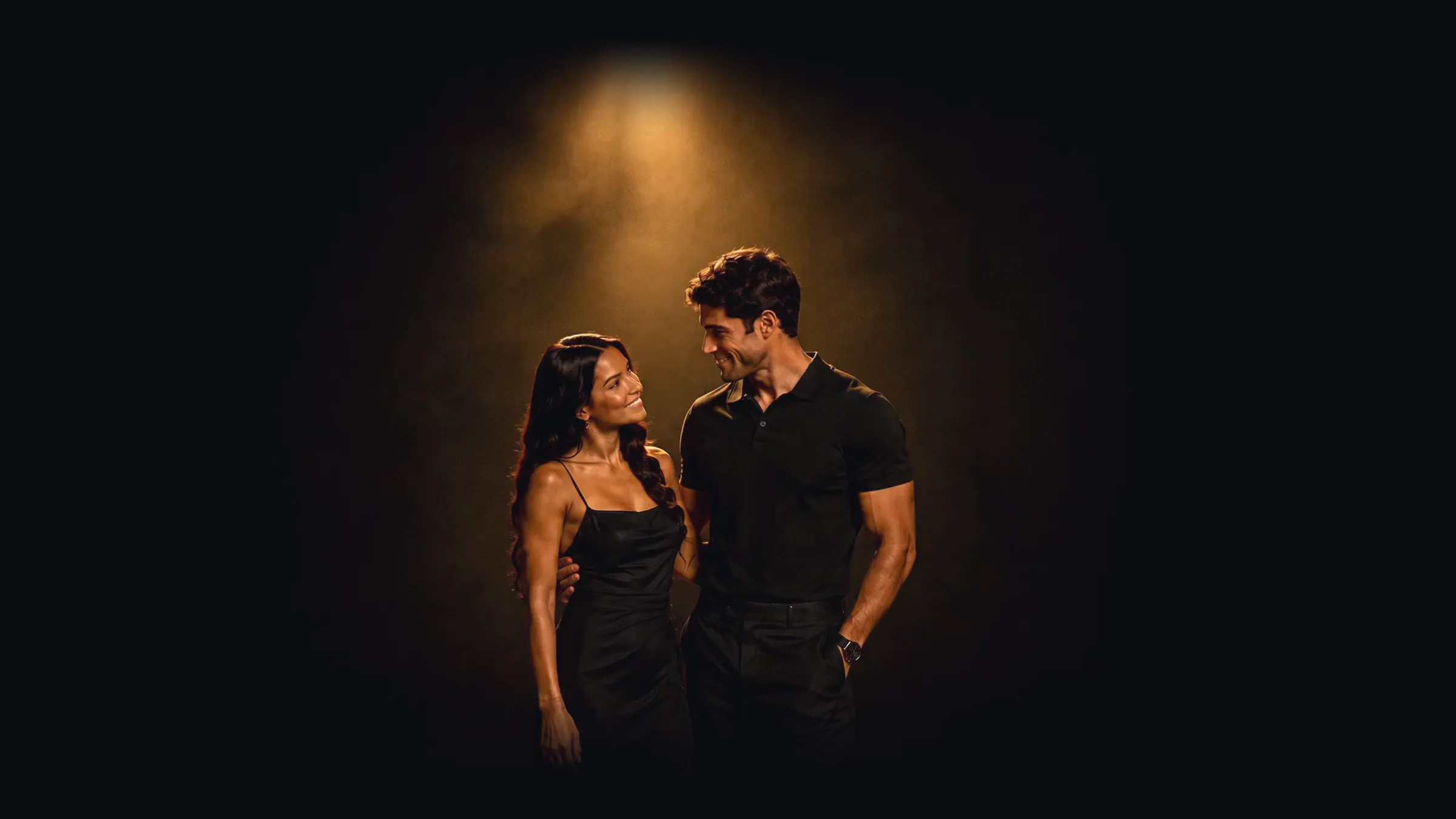 A man and a woman, both in black, standing close together under a single warm overhead light against a black ground.